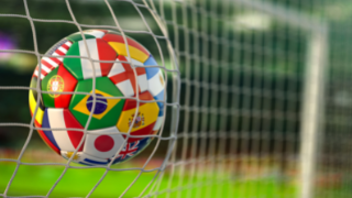 A soccer-ball with flags of several countries going into a soccer net. 